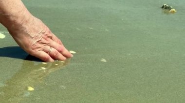 Güneşli bir sabahta, bir kadının avuç içi ıslak deniz kumları ve kıyıya yakın bir deniz dalgası ile oynar.