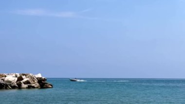 Güneşli bir günde, denizin sakin bir yüzeyinde, taştan bir tepenin arkasında bir sürat teknesi yelken açar.