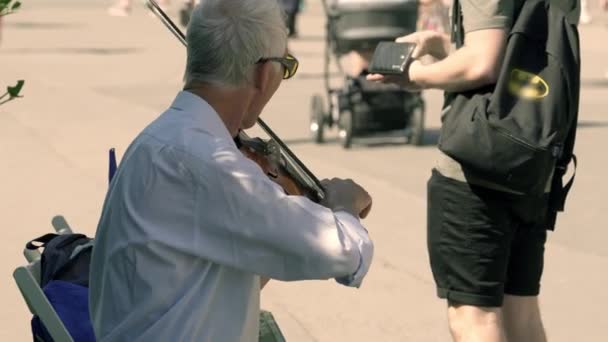 灰色の髪のウクライナの音楽家は晴れた日に街の通りでバイオリンを演奏し 若い男は財布からバイオリニストにコインを投げます — ストック動画