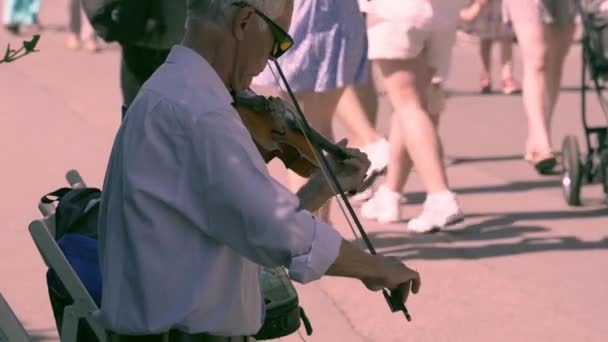 灰色の髪のウクライナの音楽家は 晴れた日に街の通りでバイオリンを演奏しています人々は通りを通ります 背景に近くを歩く小さな子供の足 — ストック動画