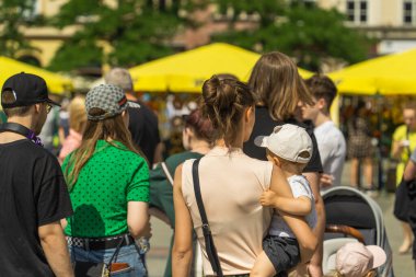 Yaz tatili elbisesi giymiş genç bir anne, yoğun bir şehir caddesi boyunca sağ elinde bir çocuğu taşır. Bir baba, güneşli bir yaz gününde başka bir çocukla birlikte yürür.