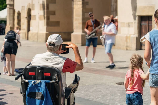 An elderly man in a wheelchair with a travel backpack uses a smartphone while relaxing in a public place of the city to shoot the performance of street musicians, next to a little girl with her mother