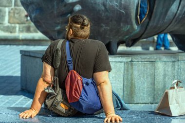 Siyah tişörtlü, sırtında iki çanta ve yanında bir kesekağıdı olan şişman bir adam şehir parkındaki mermer bir bankta demir bir kafanın arka planında oturuyor.
