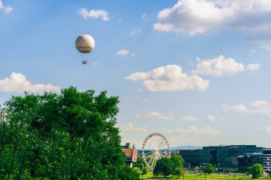 Sıcak hava balonu, dönme dolabın üstündeki mavi gökyüzünde süzülür. Ağaçlar ve şehrin sokakları güneşli bir günde insanlarla ve arabalarla doludur. Yaz şehir manzarası. Boşluğu kopyala