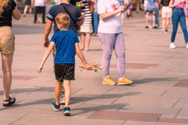 A little boy plays an airplane pilot with a toy wooden plane on a city street among adult passers-by, busy with their business and conversations, summer sunny day, street photography, children city