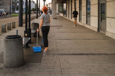 A girl in casual clothes sweeps garbage at a city tram stop in the evening, a girl with a dustpan and a broom stopped and looks dreamily into the distance, street photography, youthful dreams