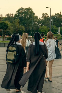 Two Polish nuns in black dresses with a backpack on their backs walk along the city street in the evening after two girls in casual summer clothes, lifestyle choice, Christian ministry, nuns in Krakow