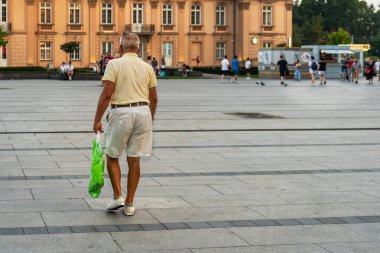 A tanned elderly lonely man in white shorts and a yellow T-shirt walks down a city street in the evening from a shopping center, shopping in a recycled plastic bag. Copy space, rear view