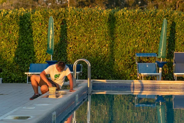 Pool cleaner, a young man in a t-shirt and shorts cleans the pool filter at sunset on a summer day in a hotel in Italy, an Italian pool cleaner at work