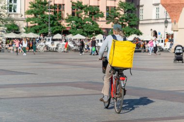 Sarı termal çantalı bir kurye, Krakow 'da güneşli bir günde eski şehrin geniş meydanındaki bir müşteriye bisikletle gider. Kurye yiyecek ve market teslimat servisi, hızlı bisiklet teslimatı.