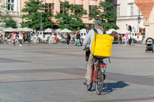 A courier with a yellow thermal bag rides a bicycle to a customer across the wide square of the old town on a sunny day in Krakow. Courier food and grocery delivery service, fast city bike delivery