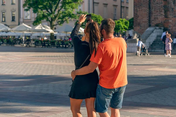 A young couple takes a selfie with a smartphone in the evening sunlight on the square of an old European city, the play of light and shadow at sunset in the city, tourist travel. Back view