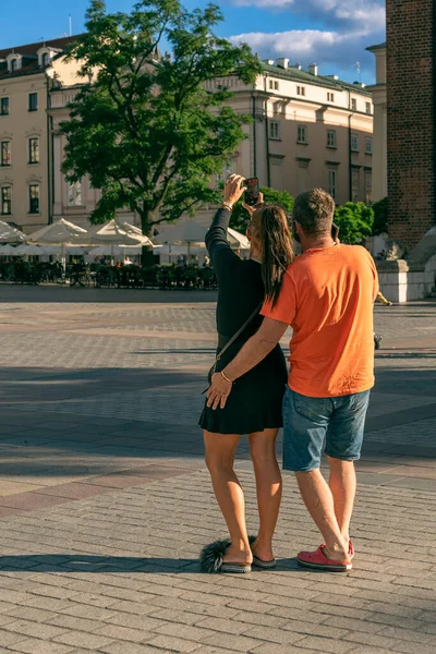 A young couple takes a selfie with a smartphone in the evening sunlight on the square of an old European city, the play of light and shadow at sunset in the city, tourist travel. Back view