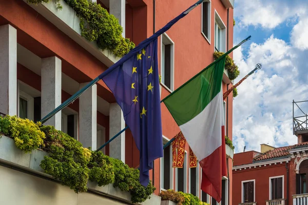 Flags of the European Union and Italy under the open sky on the balcony of an old building in Venice on a cloudy morning, the Italian flag and the European flag side by side, close-up, side view