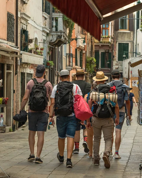 A group of tourists in hiking clothes with backpacks on the old narrow street of Venice on a summer morning, friends tourist travel, historical sights of Italy, back view