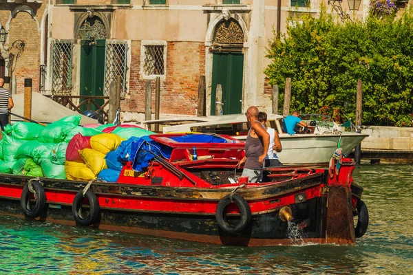 A sailor and a boy operate a cargo boat with multi-colored bags of cargo, the boat sails along the canal of the street in Venice, the transport of goods by water transport through the Venetian streets