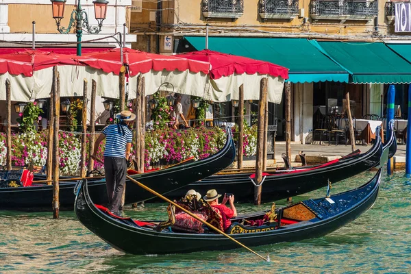 Gondolda duran bir gondolcu elinde küreği olan bir tekneyi kullanıyor. Yaşlı bir çift turist, Venedik 'teki bir caddenin kıyısındaki akıllı telefonlardan yolculuk videosu çekiyor.