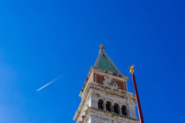 İtalya, Venedik 'teki St. Mark Meydanı' ndaki çan kulesinin tepesinde bir jet uçağının uçtuğu açık bir gökyüzüne çıkıyor. Çan kulesinin tepesinde bir işçi figürü var.