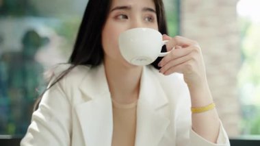 Güzel Asyalı kadın bir kafede rahatça kahvesini yudumluyor. Krema dudaklarına köpürüp kahve kokusunu alana kadar yudumluyor..