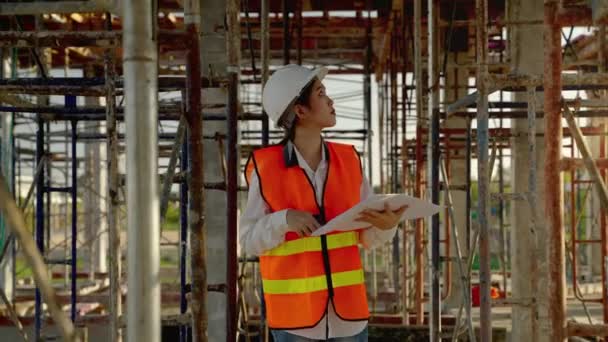 設計図を持つ若いアジアの女性エンジニアは 建設計画に従って動作するかどうかを確認するために歩いているかどうか建設現場で — ストック動画
