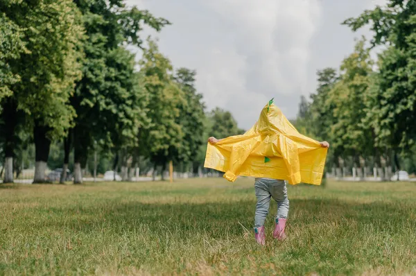 Gadis kecil yang bahagia dengan jas hujan kuning dan sepatu karet berlumpur berlari di jalan tanah melalui rumput hijau dekat ladang benih pemerkosaan yang mekar memegang badak di tangan. Masa kecil yang riang. — Stok Foto