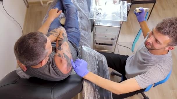 Tattoo artist fills a tattoo on the shoulder of a person with a tattoo machine — Wideo stockowe