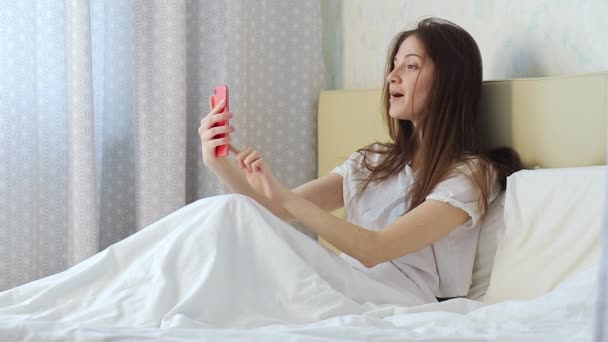 Молода дівчина тримає телефон в руках і розмовляє через відеодзвінок з другом в ліжку — стокове відео