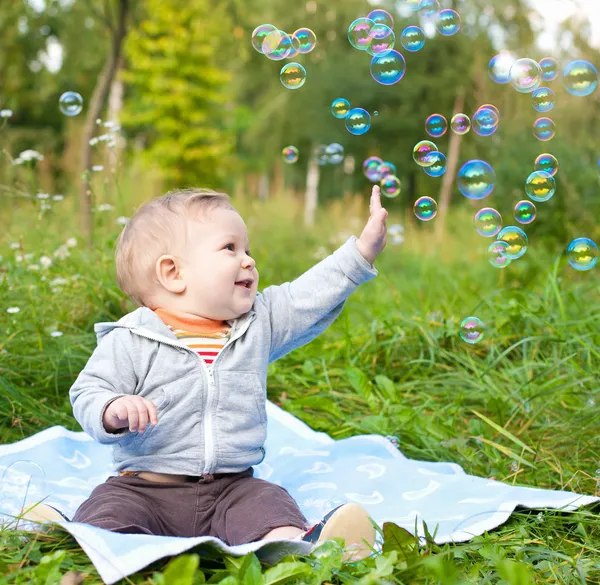 Хлопчик сидить на зеленій траві, граючи з мильними бульбашками Стокове Фото