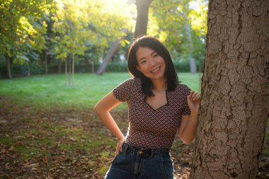 Genç mutlu ve güzel Asyalı bir kadının dış mekan portresi. Sonbaharda şehir parkında, yerde yapraklarla dolu rahat ve neşeli bir şekilde eğleniyor.