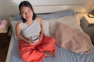 lifestyle home portrait of young happy and beautiful Asian Japanese woman pregnant sitting on bed holding ultrasound photo excited in pregnancy and maternity concept