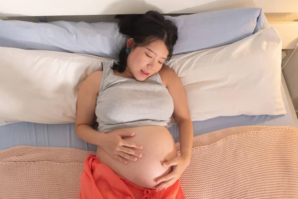 lifestyle home portrait of young happy and beautiful Asian Chinese woman pregnant lying relaxed on bed excited about maternity touching her belly in pregnancy concept