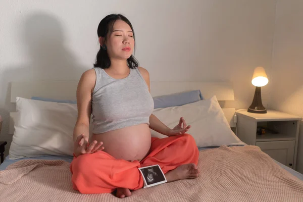 lifestyle home portrait of young happy and beautiful Asian Japanese woman pregnant doing relaxing yoga exercise on bed in maternity and pregnancy concept