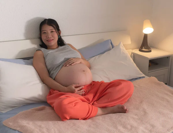 lifestyle home portrait of young happy and beautiful Asian Japanese woman pregnant lying relaxed on bed excited about maternity touching her belly in pregnancy concept