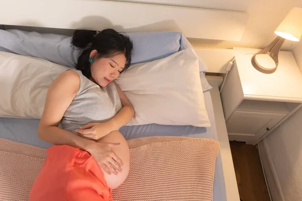 lifestyle home portrait of young happy and beautiful Asian Japanese woman pregnant lying relaxed on bed excited about maternity touching her belly in pregnancy concept