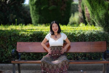 Hamile, mutlu ve güzel Asyalı bir Çinli kadın karnını gösteriyor. Parktaki bankta mutlu bir şekilde gülümsüyor. Hamilelik ve hamilelik konseptiyle.