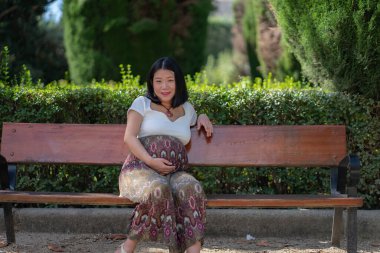 Hamile, mutlu ve güzel Asyalı bir Çinli kadın karnını gösteriyor. Parktaki bankta mutlu bir şekilde gülümsüyor. Hamilelik ve hamilelik konseptiyle.