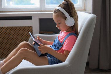 Gülümseyen küçük kız kanepede oturmuş online oyunlar oynuyor, internette sörf yapıyor, tablet üzerindeki komik uygulamaları kullanıyor.