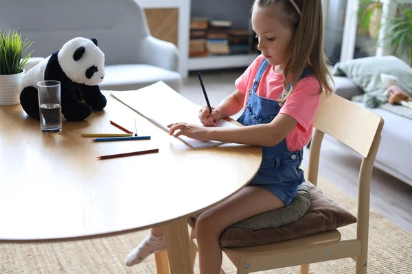 与一只玩具熊猫熊一起坐在桌旁的快乐女孩一边享受着创造性的活动 一边用铅笔在相册中着色 — 图库照片