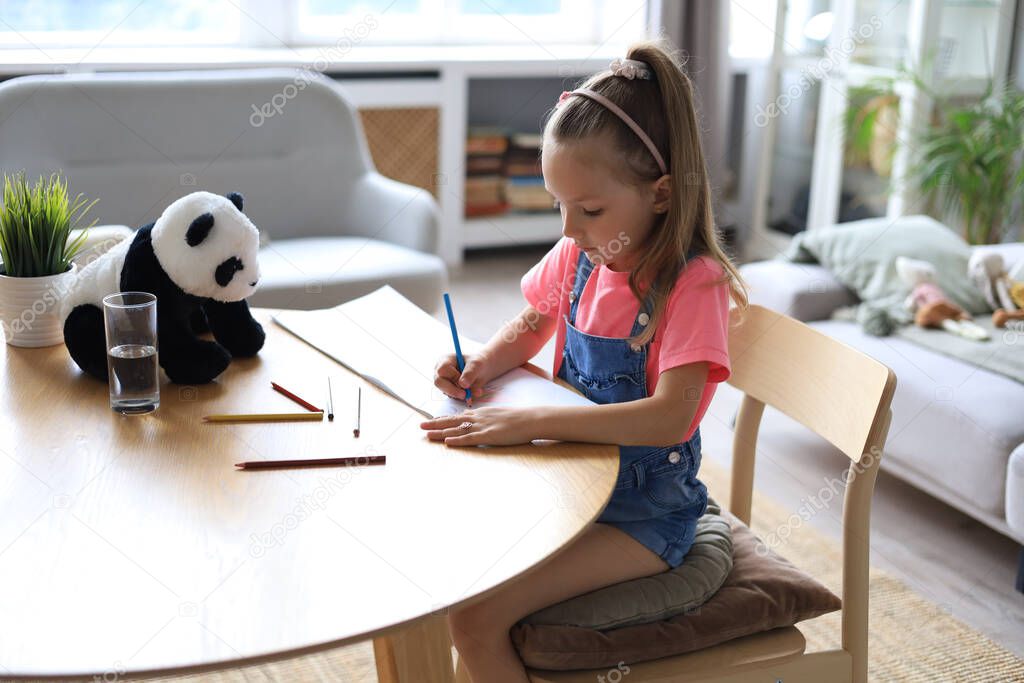 Smilling happy girl sitting in the table with a toy panda bear near to her enjoying creative activity, drawing pencils coloring pictures in albums