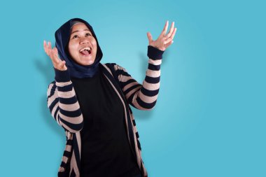 Mutlu Asyalı Müslüman kadın ellerini kaldırıp mavi arka plandan sürpriz bir ödül bekliyor.