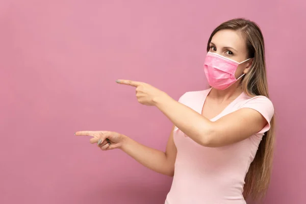 Mujer rubia en camiseta rosa y mascarilla apunta al espacio de texto sobre la pared rosa — Foto de Stock