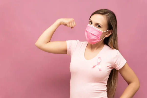 Mujer muestra fuerza como símbolo de la lucha contra el cáncer de mama en frente de fondo rosa y el uso de mascarilla — Foto de Stock
