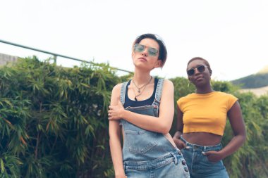 Farklı ırktan iki kadın kameranın önünde poz veriyor. Arka planda doğa var.