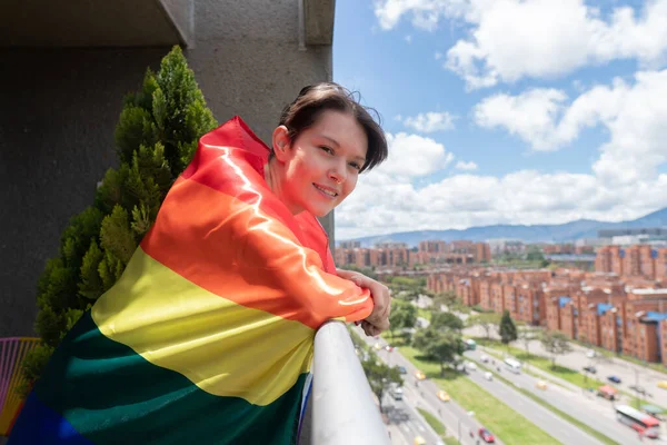Young gay woman looking from her balcony proudly wearing the LGBT flag on a sunny day — ストック写真