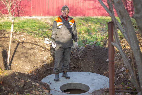 Výstavba septické nádrže pro venkovský dům. Pracovník v montérkách stojí na betonových kruzích septické nádrže s poklopem — Stock fotografie