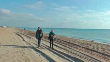 Bir adam ve bir kadın güneşli bir sonbahar gününde kumlu sahil boyunca yürür ve gülümser.