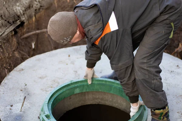 Ein Mann installiert einen Kanalschacht an einem Klärbecken aus Betonringen. Bau von Kanalnetzen im Dorf lizenzfreie Stockfotos