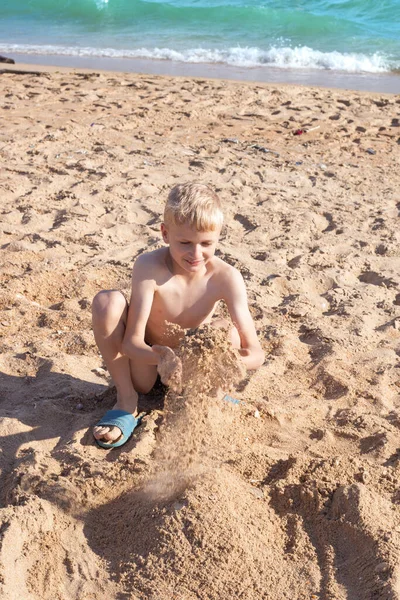 금발의 아이가 모래를 가지고 놀다가 모래를 버린다 여름날 해변에서의 — 스톡 사진