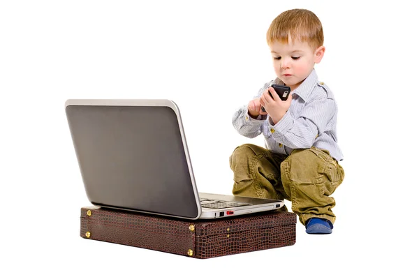 Lindo niño pequeño marca en un teléfono móvil mientras está sentado al lado de la computadora portátil — Foto de Stock