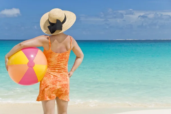 Žena v oranžové šaty a slaměný klobouk, nesoucí míč beach na pláži — Stock fotografie